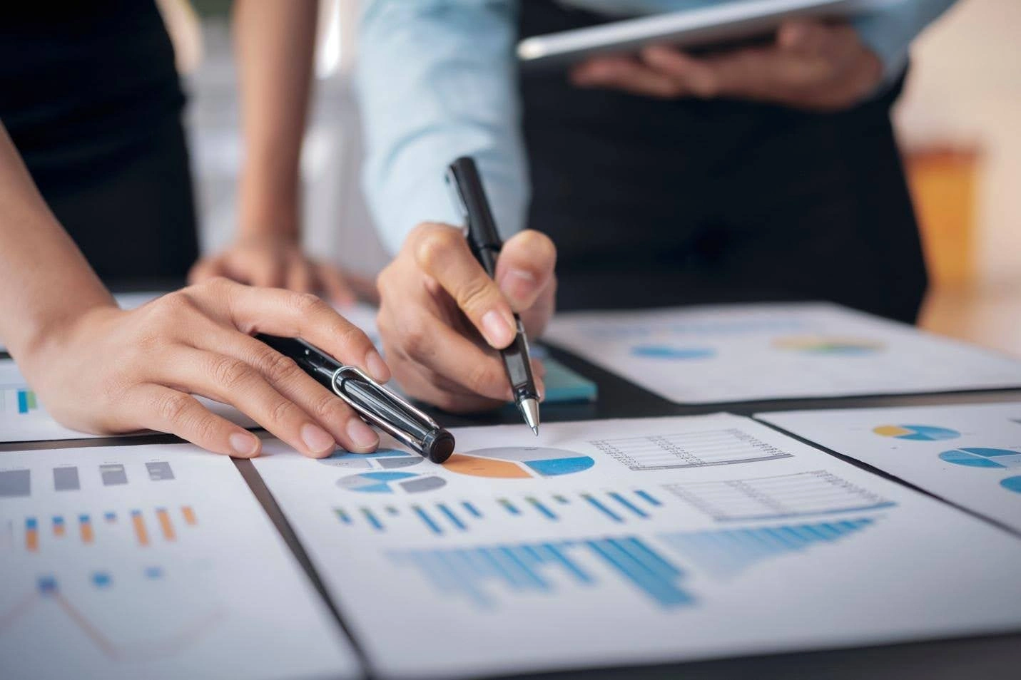 A close-up shot of two people's hands pointing with pens at various printed documents, which display pie charts and bar graphs, during a data analysis meeting.